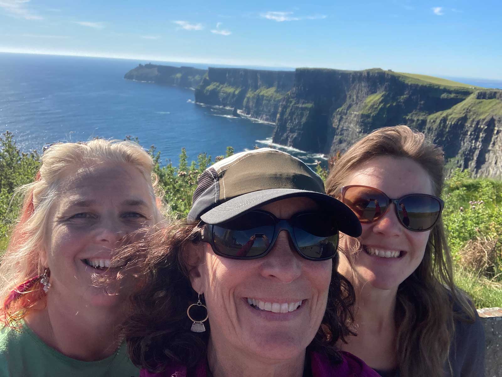 Closeup of three women's faces with the dramatic Irish coastal cliffs in the background, on a sunny day